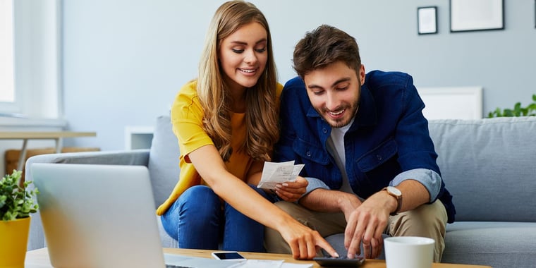 Happy young couple paying bills together and managing budget, sitting on the sofa and using calculator and laptop